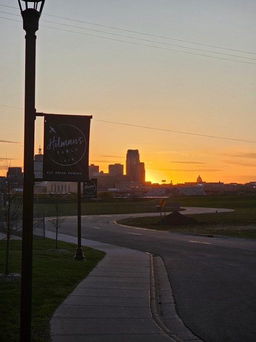 Sunset over a city skyline with a sign for "Holman's Table" in the foreground, casting a warm glow.