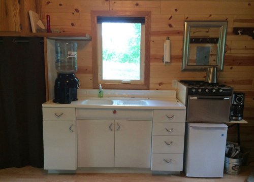 A rustic kitchen area with a sink, water cooler, small fridge, and a window overlooking greenery.