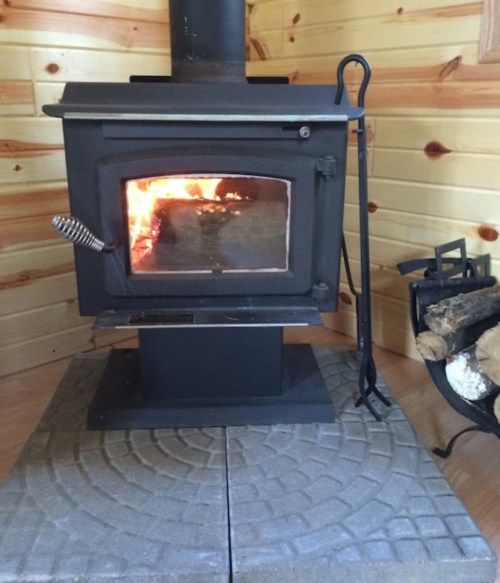 A black wood stove with a visible fire inside, set on a stone base, next to a log holder in a wooden room.