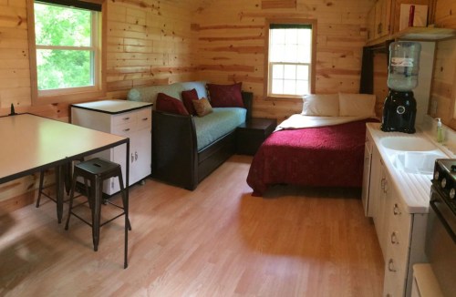 Cozy cabin interior featuring a bed, sofa, dining table, and kitchenette with wooden walls and natural light.