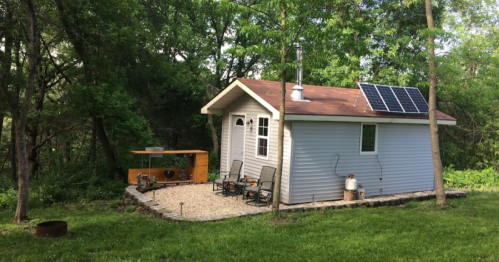 A small, light blue cabin with a solar panel, surrounded by greenery and featuring outdoor seating and a gravel area.