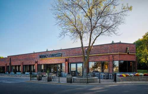 Exterior view of a brick restaurant with large windows, a tree in front, and colorful flowers along the walkway.