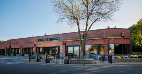 Exterior view of a brick restaurant with large windows, a tree, and colorful flowers in front. Clear blue sky above.