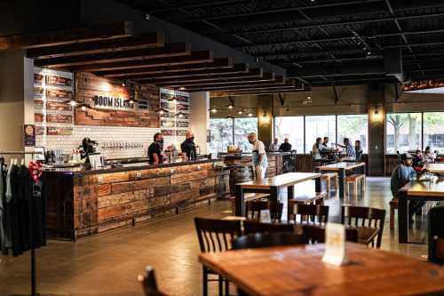 A spacious, modern bar with wooden decor, patrons seated at tables, and staff serving drinks at the counter.