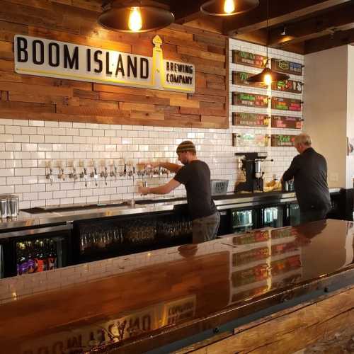 Two staff members serve drinks at the Boom Island Brewing Company bar, featuring a wooden wall and multiple beer taps.