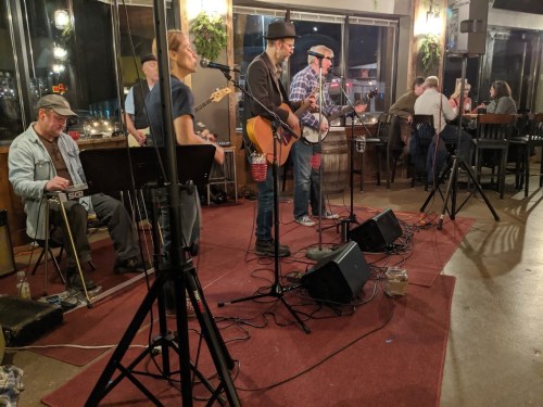 A live band performs in a cozy venue, with musicians playing guitar and banjo, while an audience enjoys the music.
