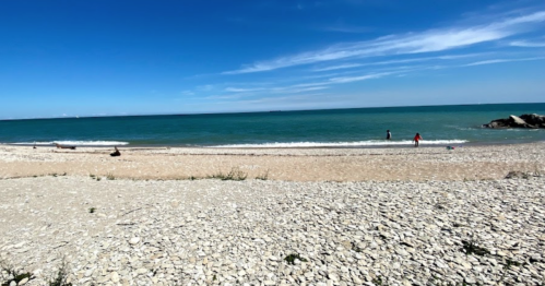 A serene beach scene with pebbles, gentle waves, and two people enjoying the water under a clear blue sky.