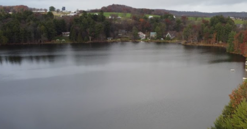 A serene lake surrounded by trees and houses, reflecting the cloudy sky and autumn colors.