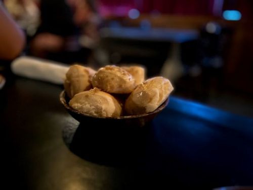 A small bowl of golden-brown bread rolls sits on a dark table, with a blurred background.