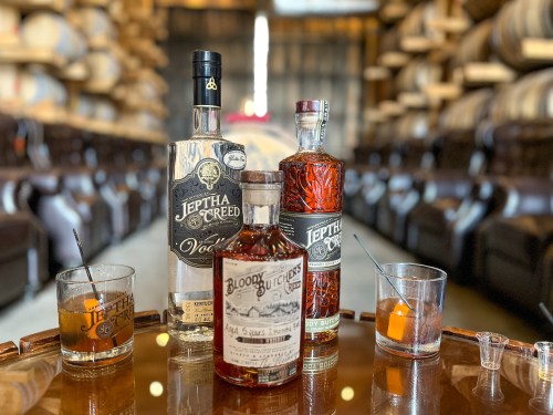 Three whiskey bottles on a wooden table, with glasses and a backdrop of stacked barrels in a distillery.