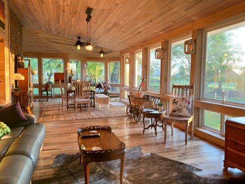 Cozy sunlit room with wooden walls, a mix of seating, and large windows overlooking a green landscape.
