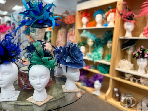 A display of colorful hats and fascinators on mannequin heads in a boutique, with shelves of accessories in the background.