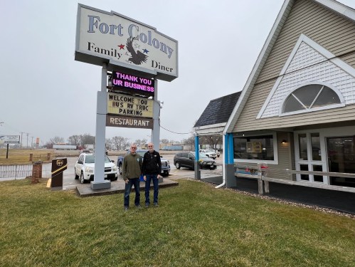 Two men stand outside Fort Colony Family Diner, with a sign thanking customers and indicating bus and truck parking.