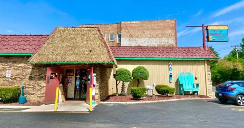 A colorful restaurant with a thatched roof, surrounded by greenery and a blue sky, featuring a sign and outdoor seating.