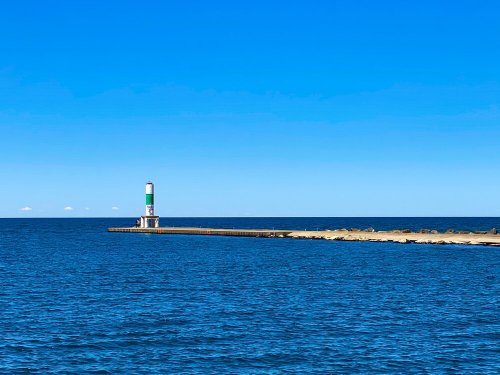 A green and white lighthouse stands on a pier extending into calm blue waters under a clear blue sky.