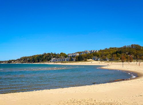 A serene beach scene with golden sand, gentle waves, and a clear blue sky, lined by green hills and buildings in the distance.