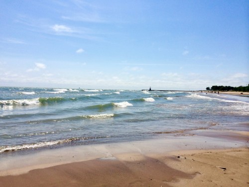 A serene beach scene with gentle waves, a clear blue sky, and a distant lighthouse on the horizon.