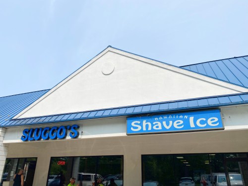 Sign for Sluggo's Hawaiian Shave Ice, featuring blue lettering on a light-colored building with a blue roof.