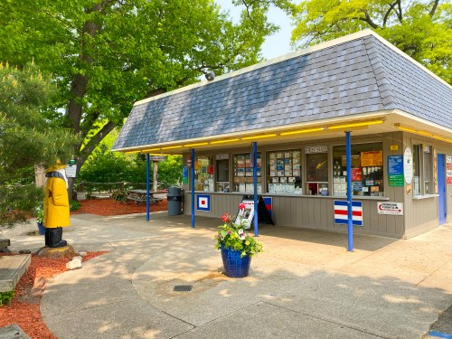 A small, colorful building with a blue roof, surrounded by trees and a picnic area, featuring a wooden statue nearby.