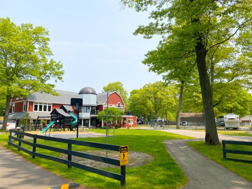 A playground with a slide and climbing structure near a red building, surrounded by green trees and a walking path.