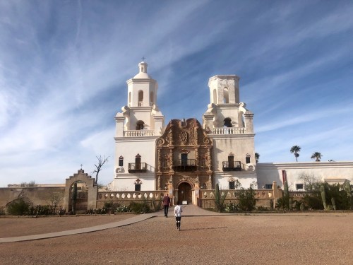 A historic building with two towers and ornate architecture, set against a blue sky, with a person walking towards it.