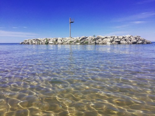 A rocky island stands in calm, clear water under a bright blue sky, with a tall structure visible on the island.