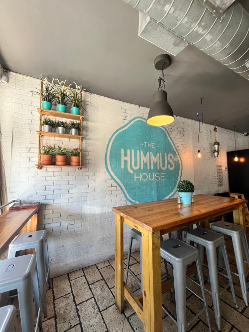 Cozy interior of "The Hummus House" with wooden tables, potted plants, and a bright mural on a white brick wall.