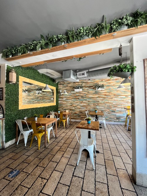 A cozy restaurant interior with green plants, wooden tables, and a decorative mirror on a textured wall.
