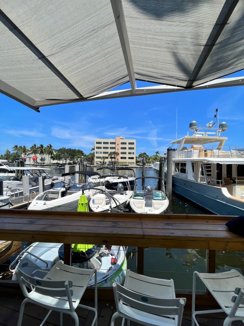 View of a marina with boats docked, a building in the background, and a shaded seating area in the foreground.