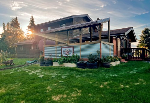 A cozy house with a deck, surrounded by greenery and flower beds, under a sunset sky.