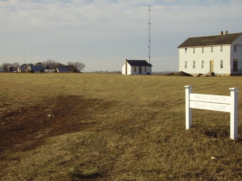 A grassy area with a sign reading "French Iotarian Colony Village" and several buildings in the background under a cloudy sky.