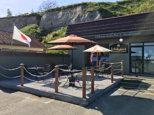 Exterior of Ichikawa Sushi Bar & Steakhouse with outdoor seating, umbrellas, and a Japanese flag. Green hillside in background.