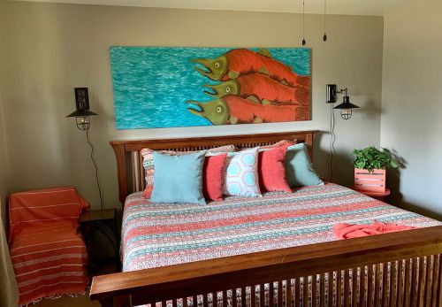 A cozy bedroom featuring a wooden bed with colorful pillows, a fish-themed painting, and a small side table.