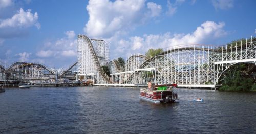 A boat floats on a river near a wooden roller coaster under a blue sky with fluffy clouds.