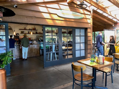 A cozy café with wooden decor, featuring patrons at the counter and tables, surrounded by greenery.