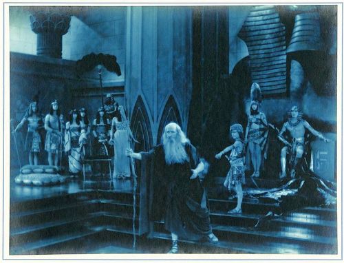 A dramatic scene featuring a bearded figure in ancient attire, surrounded by characters in a blue-toned, theatrical setting.