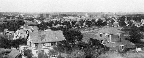 Black and white panoramic view of a town with houses, trees, and distant buildings under a clear sky.