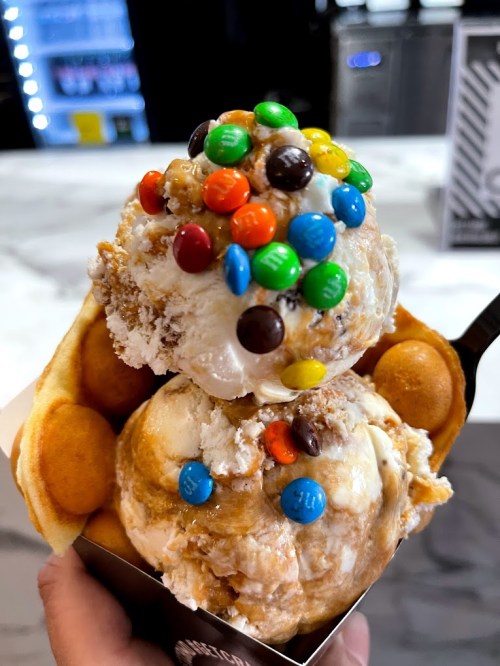 A close-up of a waffle bowl filled with two scoops of ice cream topped with colorful candy and crunchy bits.