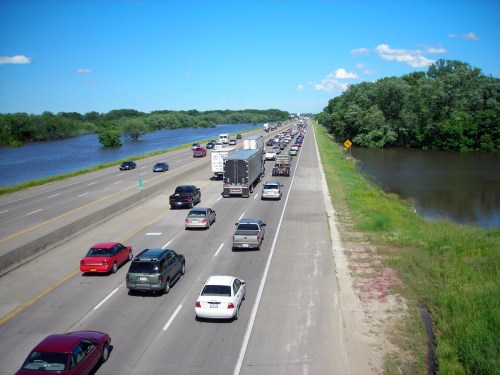Traffic jam on a highway beside a river, with lush greenery and blue skies in the background.