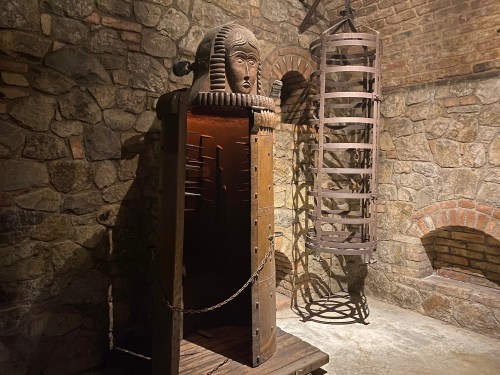 A dimly lit medieval torture chamber featuring a spiked iron maiden and a circular torture device against a stone wall.