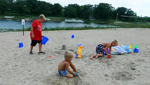 Three children play in the sand at a beach, building sandcastles and using colorful buckets near a river.