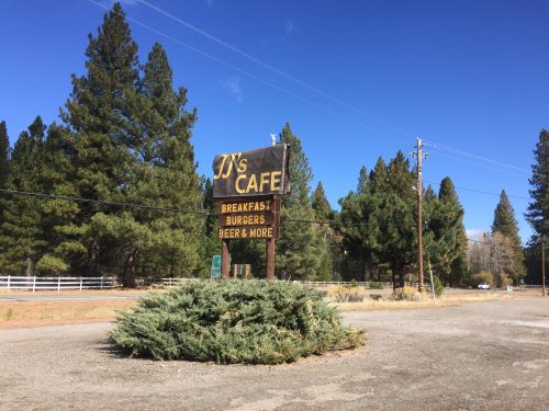 Sign for J's Cafe featuring breakfast, burgers, and beer, surrounded by trees and a clear blue sky.