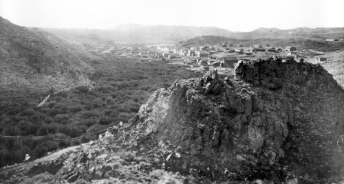 A black and white landscape showing a rocky foreground with a valley and distant town nestled among hills.