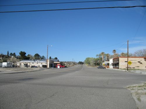 A quiet intersection in a small town, featuring a few buildings, palm trees, and a clear blue sky.