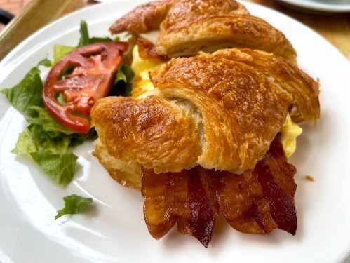 A flaky croissant sandwich filled with bacon and eggs, served with fresh lettuce and tomato slices on the side.