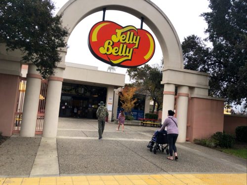 A family approaches the Jelly Belly factory entrance, with a colorful sign overhead and trees in the background.