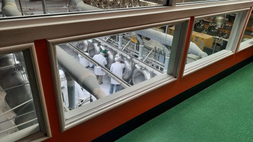 View through a window showing workers in white coats operating machinery in a factory setting.