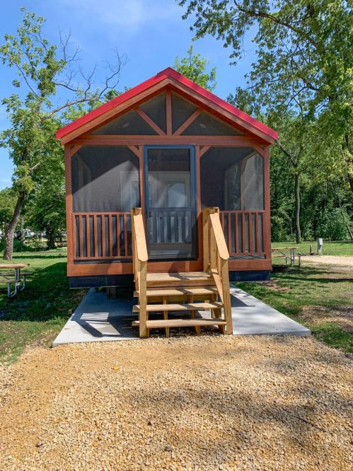 A small cabin with a red roof and screened porch, surrounded by trees and gravel pathways.