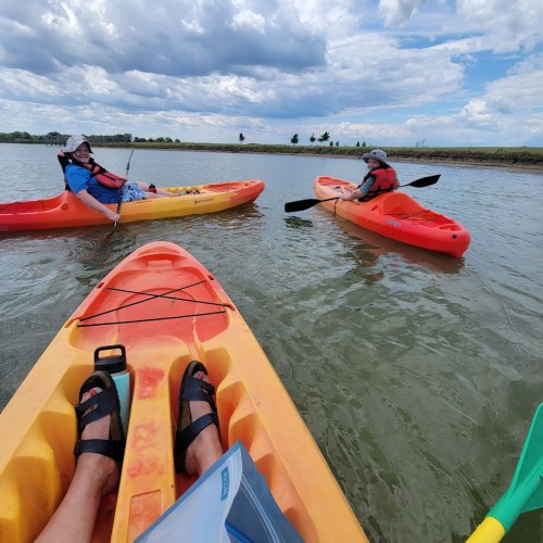 Two people kayaking on a calm waterway under a partly cloudy sky, with green banks in the background.