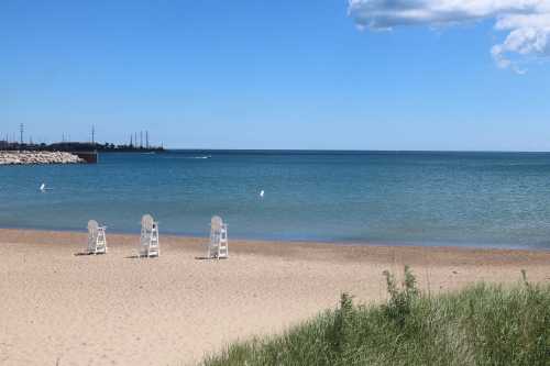 Four white lifeguard chairs on a sandy beach with calm blue water and a clear sky in the background.